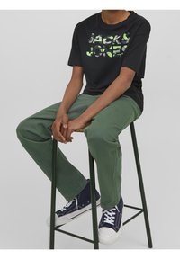 Czarna koszulka z zielonym napisem "JACK & JONES", zestawiona z zielonymi spodniami i granatowymi sneakersami z białą podeszwą, siedząca na stołku.