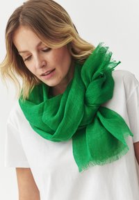 Sciarpa in lino verde con una finitura testurizzata, drappeggiata in modo leggero attorno al collo. Presenta bordi sfrangiati e un design morbido e arioso.
