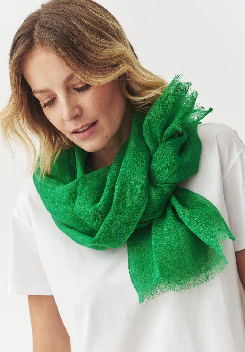 Sciarpa in lino verde con una finitura testurizzata, drappeggiata in modo leggero attorno al collo. Presenta bordi sfrangiati e un design morbido e arioso.