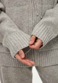Grijze gebreide sweatshirt met geribde manchetten en een rits. De stof heeft een zachte textuur en vertoont een subtiel patroon. De handen worden bij de zoom gepositioneerd.