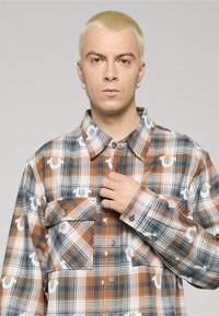 Camicia a quadri marrone e blu navy con bottoni, caratterizzata da accenti grafici bianchi, maniche lunghe, due tasche sul petto e colletto appuntito.