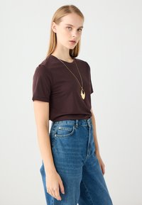 Krótki rękaw, brązowy T-shirt z miękkiego materiału noszony z jasnoniebieskimi, wysokimi dżinsami. Stylizacja uzupełniona złotym naszyjnikiem z wisiorkiem.