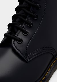 Gros plan d'une botte en cuir noire avec des lacets noirs, des œillets métalliques, des coutures détaillées et un bord de semelle cousu avec du fil jaune.