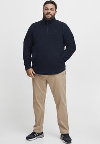 Pullover in pile blu navy con colletto alto e zip. Pantaloni beige e scarpe da ginnastica scure. Texture liscia, logo discreto sul petto.