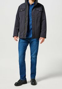 Chaqueta azul marino oscuro con cremallera y botones a presión, con dos bolsillos en el pecho, llevada sobre una camisa a cuadros azul y jeans azules, combinada con zapatos negros.