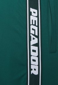 Zelené sportovní šortky s texturovaným černo-bílým vertikálním pruhem s nápisem "PECADOR". Elastický pas pro pohodlí.