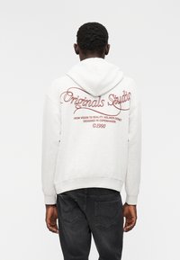 Ανοιχτό γκρι φούτερ με κουκούλα που διαθέτει κόκκινο λογότυπο και κείμενο "Originals Studio" στην πλάτη, κατασκευασμένο από μαλακό ύφασμα με άνετη εφαρμογή και ρυθμιζόμενη κουκούλα.