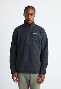 Man wearing a black fleece jacket with a zippered chest pocket and gray pants, standing against a plain white background.