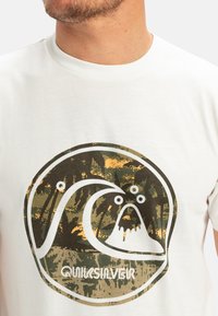 Bílá tričko s kulatým potiskem vlny a hory v zeleno-oranžovém maskáčovém vzoru, s textem "Quiksilver" dole.