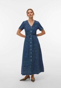 Naine seisab kätega selja taga, kandes sinist nööpidega denimmaksi kleiti ja valgel taustal sebra mustriga teravate ninadega kingi.