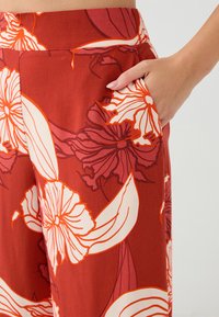 Rode bloemenpatroon broek met een gladde textuur, voorzien van een zijzak en een brede tailleband. Bloemen in crèmekleurige en oranje tinten.