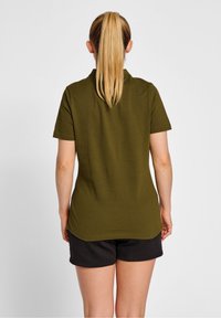 Kvinde med blond hestehale iført en olivengrøn kortærmet skjorte og sorte shorts, vendt væk mod en ensfarvet hvid baggrund.