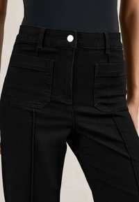 Primer plano de pantalones negros de cintura alta con bolsillos delanteros de parche, un botón plateado, pasadores para cinturón y costuras visibles.