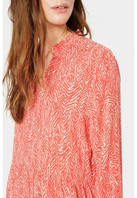 Červená blůza s vzorem a výstřihem do V a řasenými okraji. Látka má design v rybí kosti v světlejších tónech, což poskytuje texturovaný vzhled.