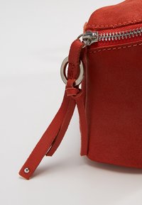 Bolso de ante rojo con cremallera plateada, con una correa de tela sujeta por un anillo de metal, textura suave y diseño minimalista.