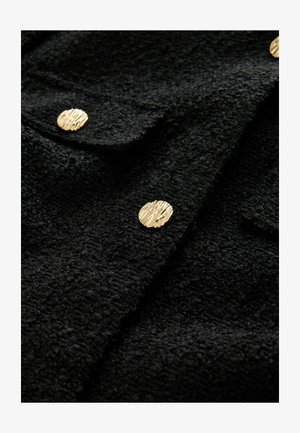 Schwarze, strukturierte Jacke mit metallisch-goldenen, runden Knöpfen mit gerilltem Muster, die kontrastierende Materialien und Details hervorheben.