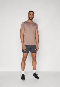 Mees seisab ühetoonilise tausta ees, seljas helesinine lühikeste varrukatega särk, tumedate mustritega lühikesed püksid, valged sokid ja mustad spordijalatsid.