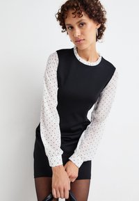 Musta mekko, jossa on tiukka vartalo-osa ja levenevät pilkulliset hihat. Kauluksessa on röyhelöreunus. Malli pitää mustaa käsilaukkua.