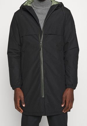 Veste noire à capuche de longueur moyenne, zippée à l'avant, portée sur un pull à col roulé côtelé gris.