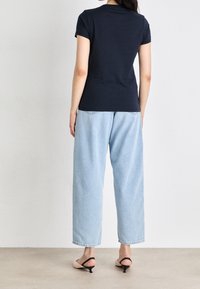 Žena ve černém tričku s krátkým rukávem, světle modrých širokých džínách a béžových sandálech na nízkém podpatku, stojící čelem ke bílé zdi.