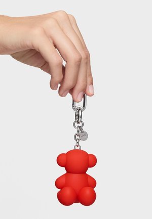 Hand hält einen silbernen Schlüsselanhänger mit einem roten Gummibärchen-Anhänger vor einem schlichten weißen Hintergrund.