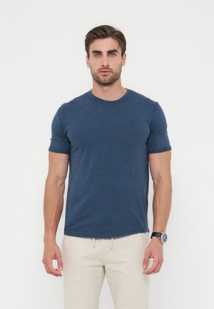 Homme aux cheveux bruns courts portant un T-shirt bleu marine à manches courtes et un pantalon beige, debout sur un fond blanc.