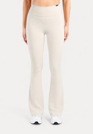 Femme portant des leggings évasés couleur crème à la taille haute avec des baskets blanches, debout devant un fond blanc uni.