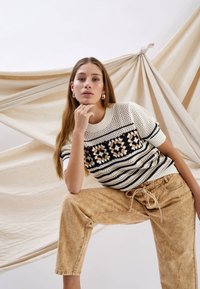 Young woman with long hair wearing a patterned knit sweater and light brown pants poses against beige fabric backdrop, resting chin on hand.