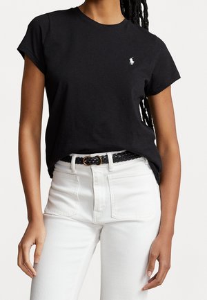 Femme portant un t-shirt noir à manches courtes avec un petit logo blanc et un pantalon blanc taille haute avec une ceinture noire tressée.