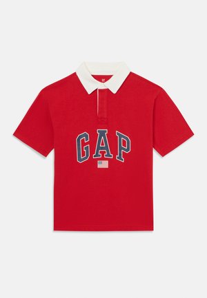 Red polo shirt made of cotton with a white collar. Features "GAP" logo in navy, with a small American flag accent below. Short sleeves.