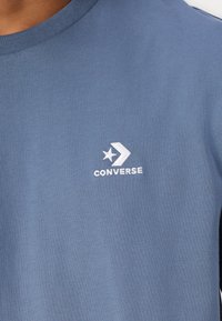 Šviesiai mėlynas medvilninis marškinėlis su apvaliomis apykaklėmis. Ant kairiojo krūtinės užrašytas mažas baltas "Converse" logotipas ir žvaigždės emblemą.