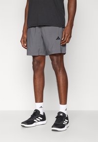 Γκρι αθλητικό σορτς με επίπεδη μέση και πλαϊνή τσέπη, συνδυασμένο με μαύρα αθλητικά παπούτσια με άσπρες ρίγες και λευκές κάλτσες.