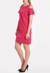 Rosa, knielanges Kleid mit kaltem Schulterdesign und Rüschenausschnitten. Glatte Textur und einfache Silhouette. Schwarze High Heels vervollständigen das Outfit.