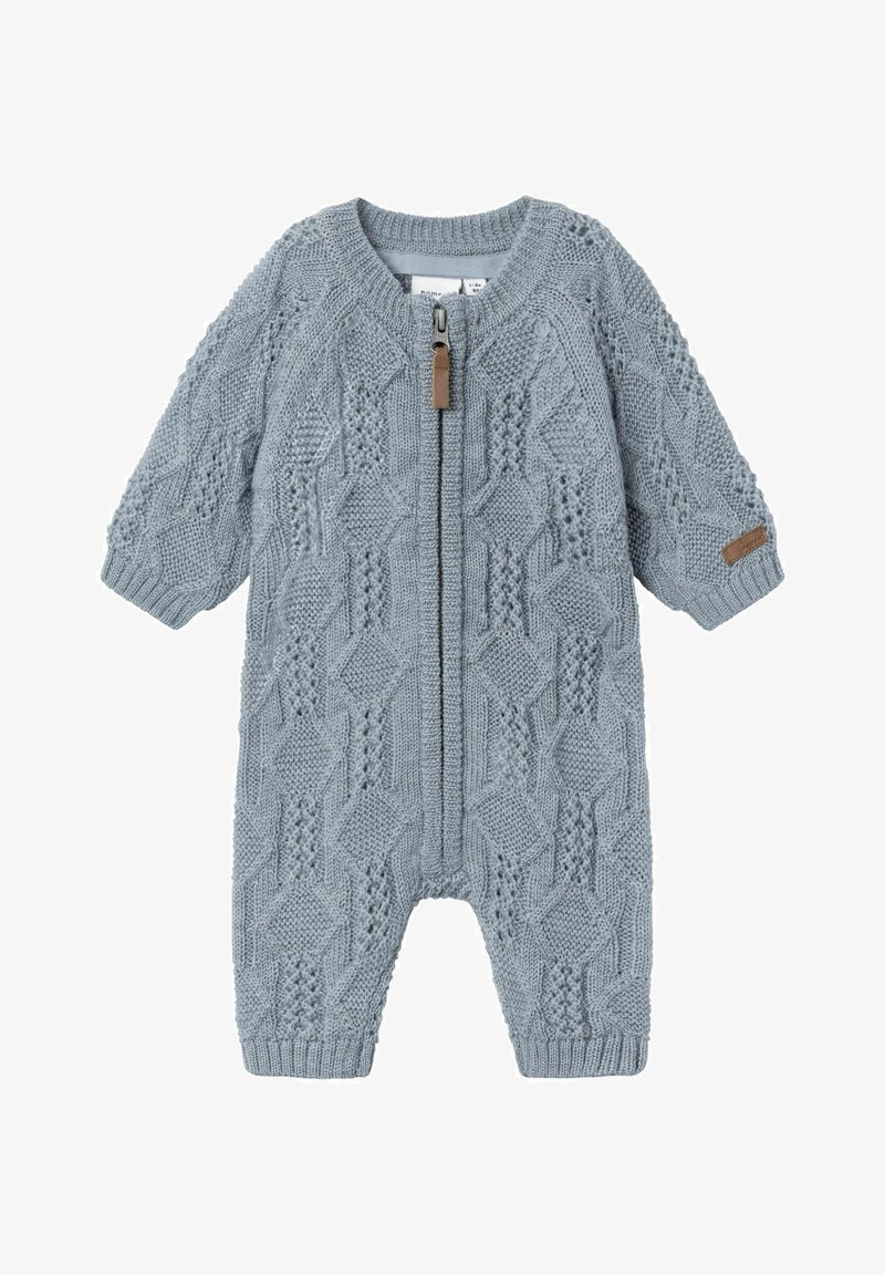 Combinaison en laine bleu tricotée avec un motif en relief, fermeture éclair sur le devant et poignets en côtes. Présente un accent en cuir beige près de la manche.