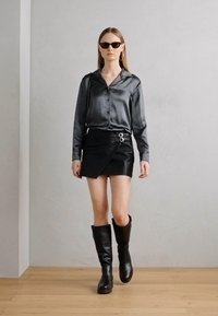 Musta satiinista pitkähihaine paita, epäsymmetrinen musta nahkaminihame hopeisilla yksityiskohdilla, polven yli ulottuvat mustat nahkasaappaat ja tummat aurinkolasit.