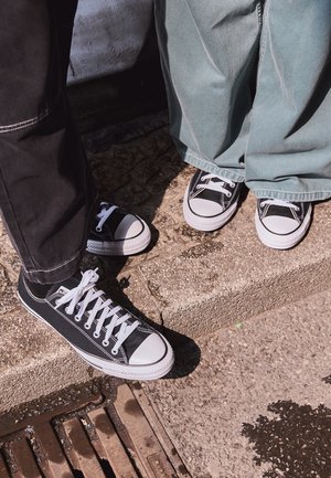 To personer iført sorte og hvide lærredssneakers står på en tekstureret fortov nær en metalrist.