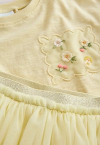 Gul kjole med broderede små blomster på bodice, bølget kant og glitrende bælte over en rynket tylskørt.