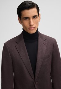 Blazer bordeaux con rever a punta, tessuto strutturato e tasca sul petto, indossato sopra un maglione a collo alto nero.