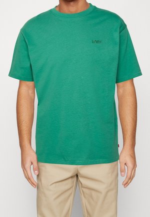 Homme portant un T-shirt à manches courtes Levi's vert et un pantalon beige, debout les bras détendus le long du corps devant un fond uni.
