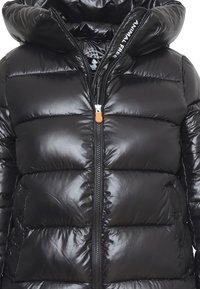 Giacca imbottita nera con colletto alto e chiusura con zip. Presenta una trama lucida, bottoni laterali e un accento arancione sulla zip.