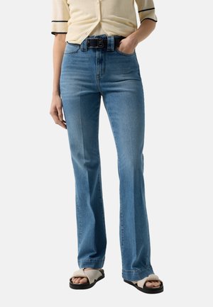 Frau trägt hohe, gerade geschnittene blaue Jeans mit schwarzem Gürtel, beige Kurzarmoberteil und beigen Sandalen mit breiten, sich kreuzenden Riemen.