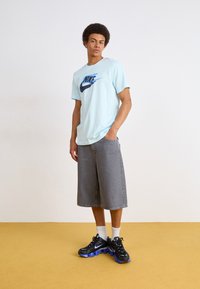 Světle modré tričko Nike má velké modré logo, ve spojení se šedými šortkami po kolena a černými botami s modrými akcenty.