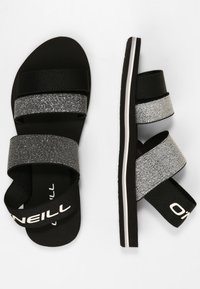 Mustat sandaalit, joissa on kaksi leveää hopeanväristä kimallevyötä, teksturoitu pohja ja musta kumipohja valkoisilla korostuksilla.