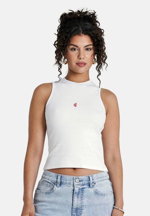Witte geribbelde tanktop met een hoge hals, voorzien van een klein rood logo op de borst. Gecombineerd met lichtblauwe high-waisted jeans.