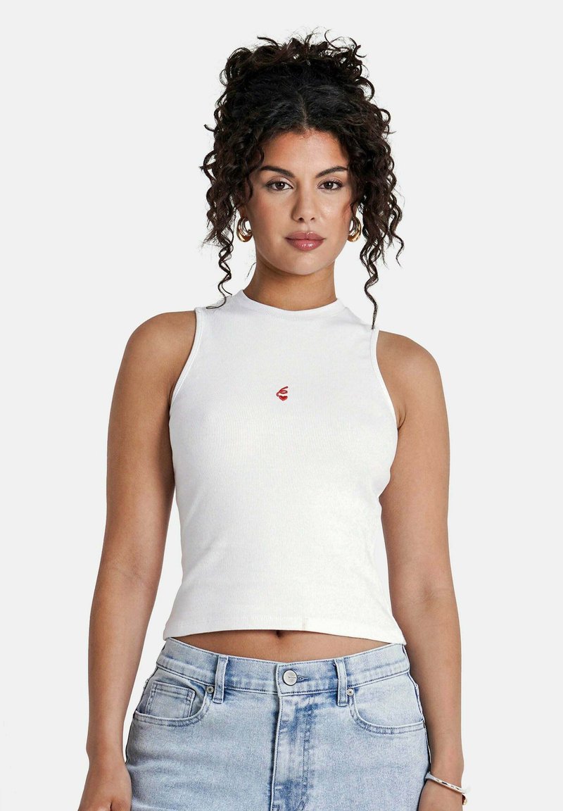 Top de tirantes blanco con textura acanalada y un cuello alto, con un pequeño logo rojo en el pecho. Combinado con jeans de tiro alto en azul claro.