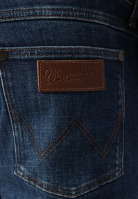 Donkerblauwe spijkerbroek van denim met een bruine leren Wrangler-label. Kenmerkt zich door oranje stiksels en een kenmerkend zigzagpatroon op het achterzak.