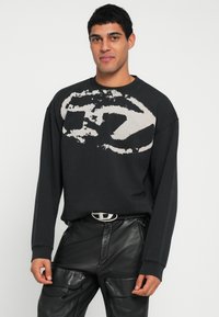 Sweatshirt preta com um grande desenho abstrato branco na frente, com gola redonda e mangas compridas. Combinada com calças pretas de couro.