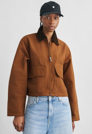 Junge Person mit schwarzer Kappe, brauner Reißverschlussjacke mit Klappentaschen und hellblauen Jeans, stehend vor einfachem Hintergrund.