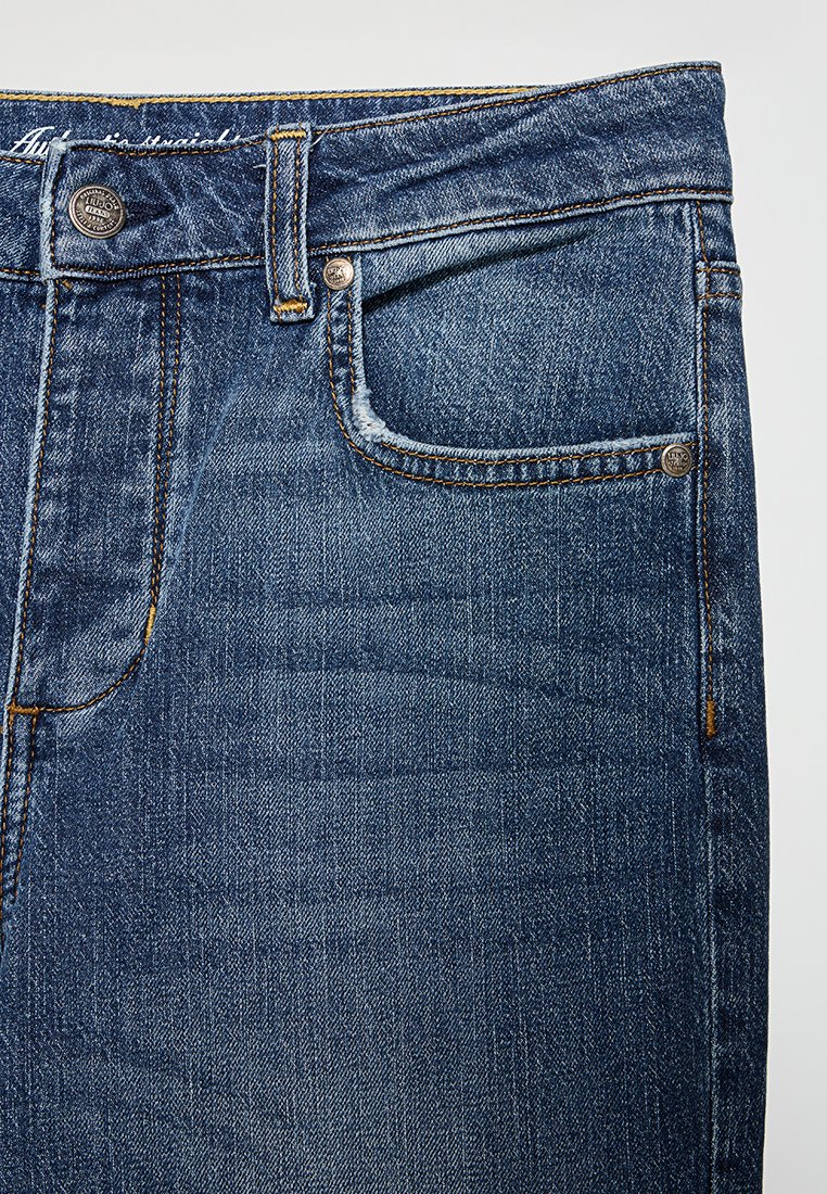 Jeans in denim blu che mostrano la parte anteriore superiore con cintura, bottone, passante per la cintura e tasca anteriore con rivetti metallici.