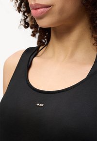 Γυναίκα που φοράει μαύρο τοπ χωρίς μανίκια με μικρό ασημένιο λογότυπο "BOSS" κεντραρισμένο κάτω από το λαιμό, με μερικά σγουρά μαλλιά ορατά.